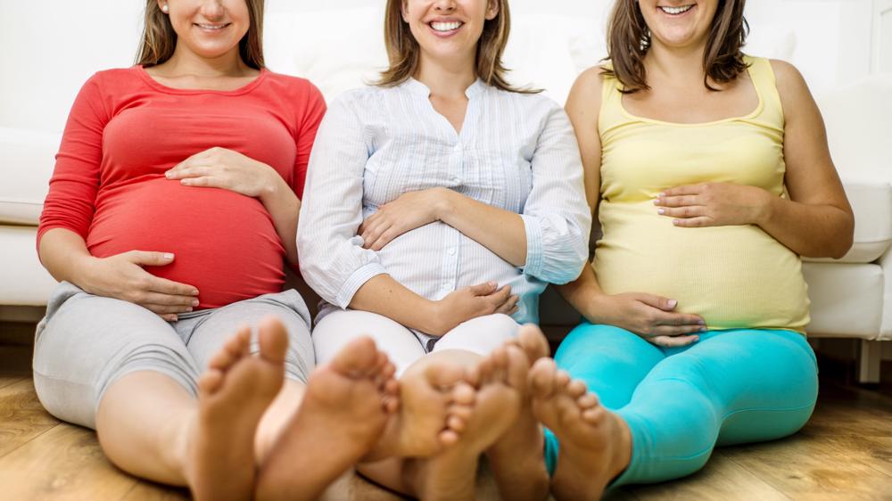 Three pregnant women are shown sitting on a floor in front of a sofa holding their stomachs